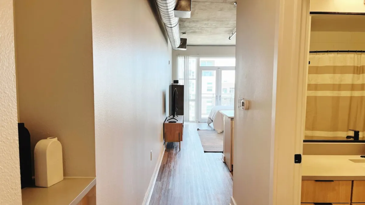 A view down a long hallway into an open living space, where sunlight streams through a large window. The scene features clean lines, contemporary furnishings, and areas for both relaxation and entertaining. The natural light and open layout evoke a welcoming, modern ambiance.