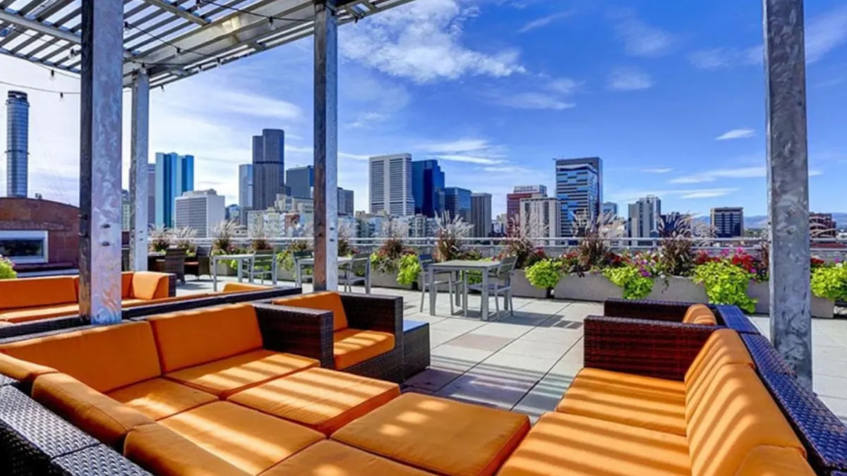 A cheerful exterior area showcases comfortable orange seating, lush planters, and sweeping views of the city skyline. The space is clearly designed for gatherings, relaxation, and enjoying the outdoors, with ample room for socializing and appreciating city sunsets.