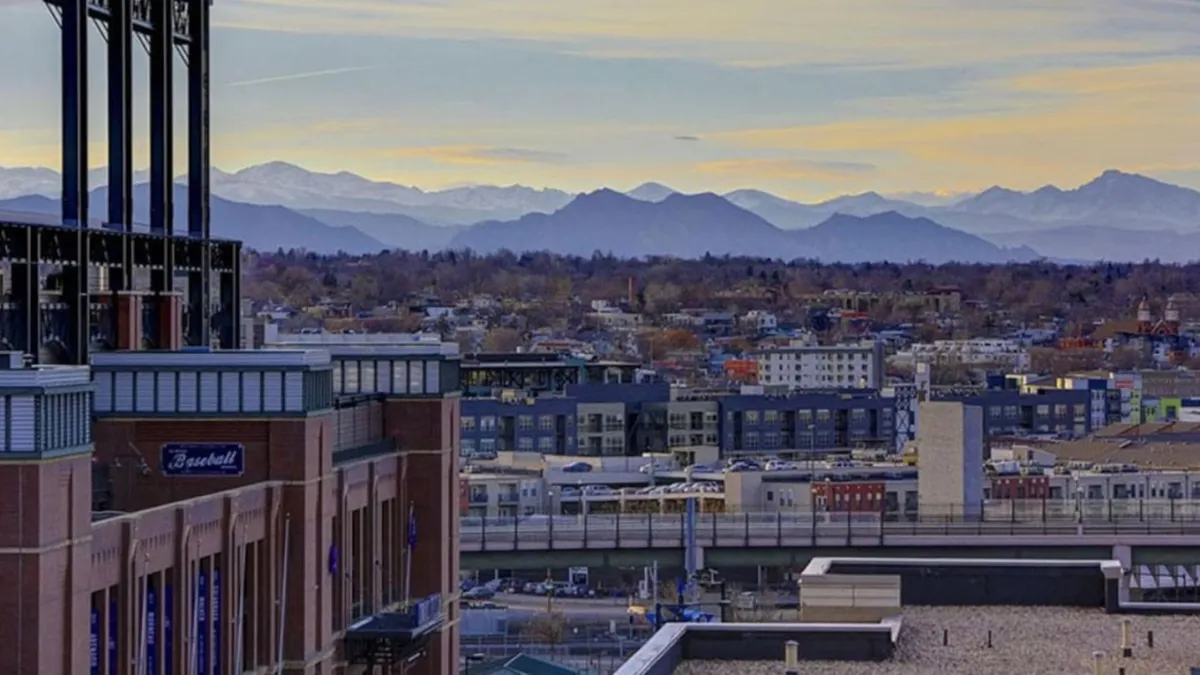 The image offers a sweeping view of Denver’s cityscape at sunset, with the iconic Coors Field stadium prominently featured in the foreground. The distant mountain range frames the urban scene, and the vibrant sky hints at both the city’s energy and its natural beauty. There are signs of seasonal festivities and hints of community gatherings around the stadium.