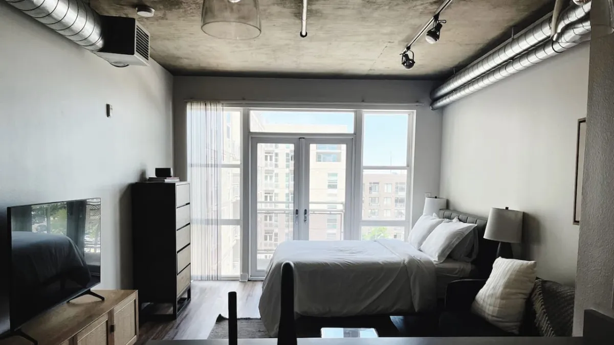 A welcoming studio sleeping area featuring a comfortable bed positioned near large windows that flood the space with natural light. A television and dresser add functionality, making the studio feel both homey and practical. The room is modern, uncluttered, and imbued with a tranquil atmosphere—ideal for relaxation after a busy day.