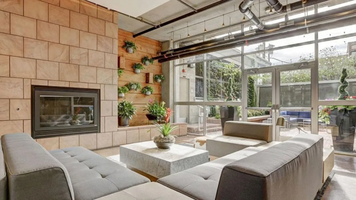 This image showcases a stylish open-plan living area centered around a modern sectional sofa and a sleek marble coffee table, with green wall planters adding touches of nature. The inviting fireplace and large glass doors contribute to a bright, welcoming atmosphere, while the setup clearly accommodates both relaxation and social gatherings.