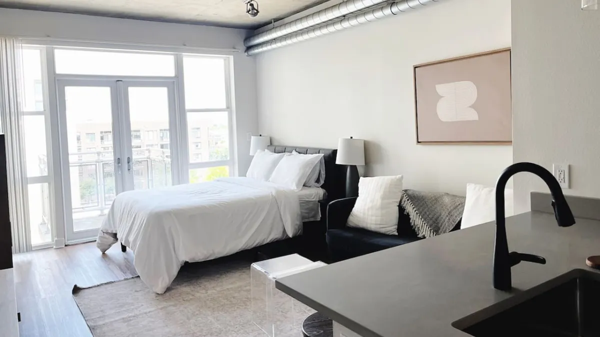 A bright, thoughtfully designed modern studio apartment featuring a cozy bed, inviting sofa, contemporary art piece, and large window letting in abundant natural light. The scene evokes a sense of calm, perfect for relaxing, reading, or sipping morning coffee while overlooking the vibrant Denver cityscape.
